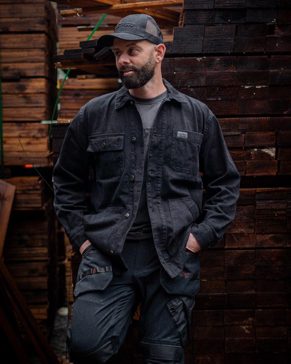 Man wearing a Black TuffStuff Cap and Black coloured TuffStuff Ranger Jacket with a lumber yard background
