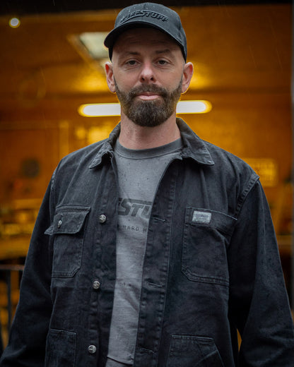Man wearing a Black TuffStuff Cap and Black coloured TuffStuff Ranger Jacket with a blurred indoor background 