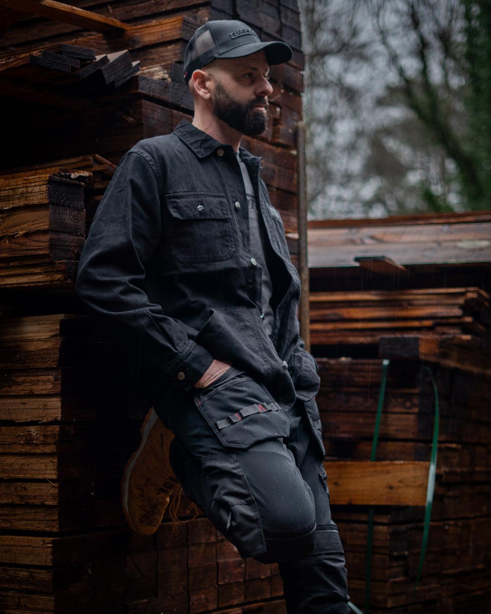 Man wearing a Black TuffStuff Cap and Black coloured TuffStuff Ranger Jacket with a lumber yard background 