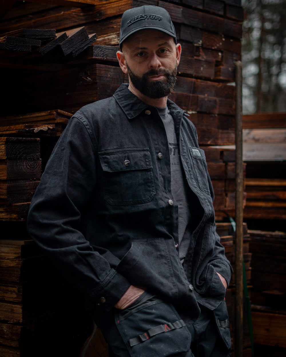 Man wearing a Black TuffStuff Cap and Black coloured TuffStuff Ranger Jacket with a lumber yard background 