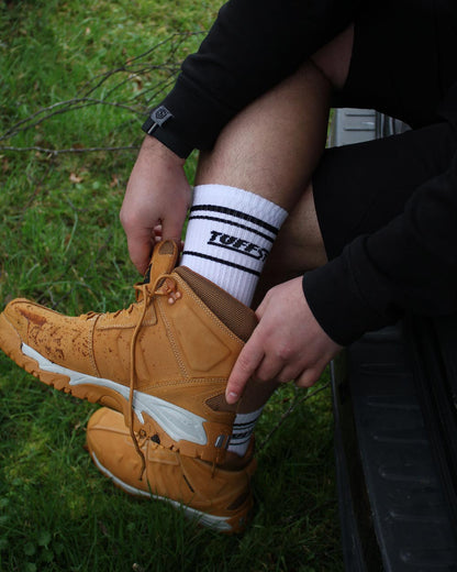 Person wearing a black jacket and white TuffStuff crew socks with black stripes, tying a brown boot outdoors on grass.